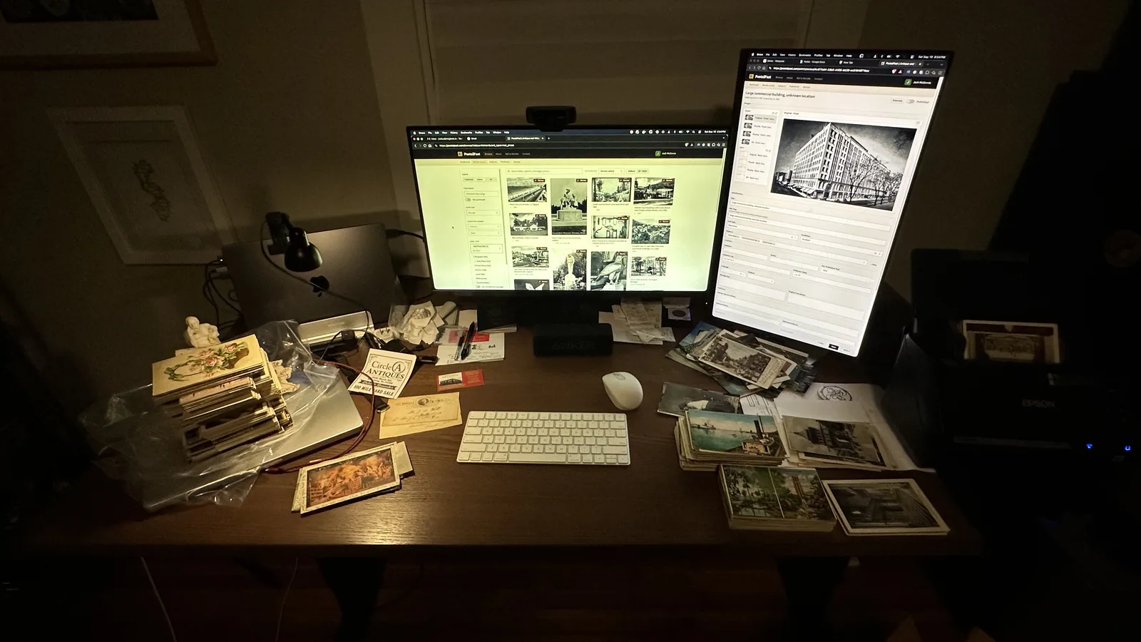 A dimly lit desk at night with monitors open to the PostalPast admin and piles of postcards waiting to be processed